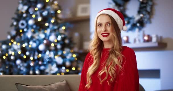 Portrét radostné bílé mladé feny v červeném svetru v santa klobouku s úsměvem na kameru ve slavnostní náladě sedí v útulném moderním pokoji se zářícím zdobeným stromem pocit štěstí. Veselé Vánoce koncept — Stock video