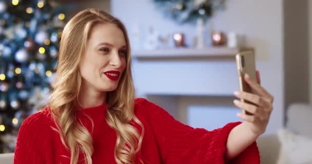 Close-up portret van Kaukasische vrolijke jonge vrouw in versierd modern huis met kerstboom spreken op video bellen op smartphone en het verzenden van de beste wensen met vakantie op kerstavond. Xmas-concept — Stockvideo