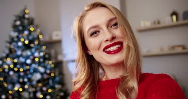 Close-up portret van blanke vrolijke jonge vrouw in versierde moderne kamer met gloeiende kerstboom videochatting en het verzenden van de beste wensen met vakantie. POV van vrouw opname van kerst video blog — Stockvideo