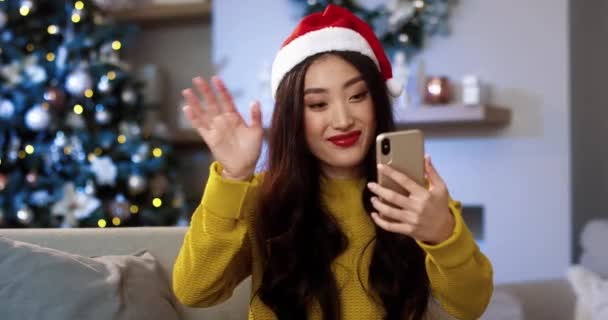 Detailní portrét asijské veselá mladá fena ve zdobeném moderním domě s zářící vánoční strom mluví na videohovoru na smartphonu a posílání přání všeho nejlepšího s dovolenou na Vánoce. Vánoční koncept — Stock video