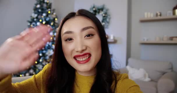 Mutlu Asyalı genç bir kadının portresini yakın çekimde, dekore edilmiş modern bir odada, Xmas ağacıyla parlayan ışıklar ile internetten konuşan ve elinde Noel hediyesi tutan. POV konsepti. İyi tatiller. — Stok video