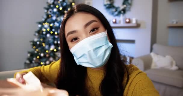 Close-up portret van gelukkige jonge Aziatische vrouw in medisch masker in kamer met gloeiende kerstboom met twinkelende lichten spreken op video online bellen en vasthouden van kerstcadeau. POV concept. quarantainevakanties — Stockvideo