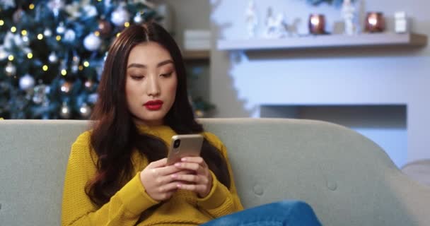 Close up portrait of joyful young beautiful Asian woman in decorated modern house with xmas tree texting on smartphone and sending greetings on Christmas. Festive mood. Xmas spirit concept — Stock Video