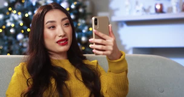 Close up portrait of cheerful young beautiful Asian woman in decorated modern house with xmas tree videochatting on smartphone and waving hand sending greetings on Christmas. Xmas spirit concept — Stock Video