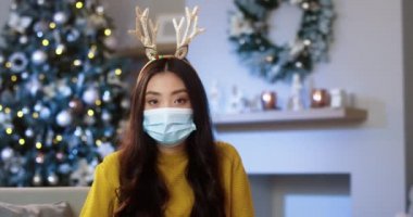 Medikal maskeli, rahat, modern bir odada oturmuş kameraya bakan Asyalı güzel bir kadının portresini yakından çek. Noel süsü. Karantinada tatil sezonu. Mutlu Noeller.
