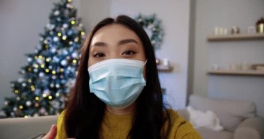Tıbbi maskeli neşeli genç Asyalı kadının arka planda parıldayan Xmas ağacıyla yeni yıl arifesinde internette video konferansında konuştuğu yakın plan. POV konsepti. Karantina tatili