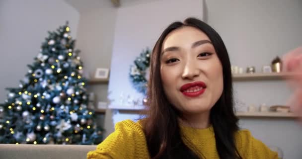초상화닫아. 화려 한 크리스마스 트리와 함께 장식 된 현대의 방에 앉아 비디오 채팅을 하며 손을 흔들고 있는 즐거운 젊은 아시아 여성 의 POV. 새해 컨셉이야. 즐거운 휴일 — 비디오