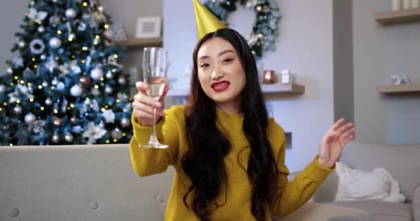 Porträtt av asiatisk glad ung vacker kvinna med glas champagne i rummet nära glödande julgran medan man talar inspelning video blogg önskar glad semester på nyår. Bloggningskoncept — Stockvideo