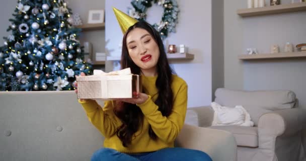 Portret van Aziatische lachende jonge vrouwelijke influencer zitten in versierde kamer in de buurt van kerstboom tijdens het spreken opname video blog wensen gelukkige feestdagen en tonen van Kerstmis aanwezig. Bloggconcept — Stockvideo