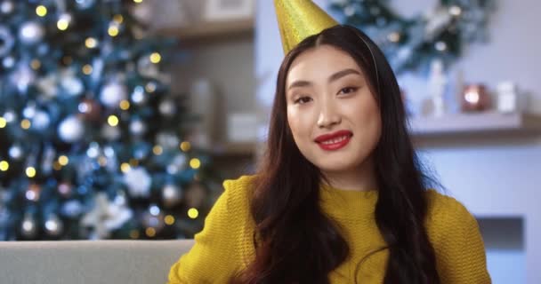 Primer plano retrato de la joven y feliz mujer asiática bonita en el ambiente festivo interior en la habitación decorada mirando a la cámara y sonriendo en la víspera de Navidad. Celebración de Año Nuevo. Vacaciones de invierno. Concepto espíritu de Navidad — Vídeo de stock