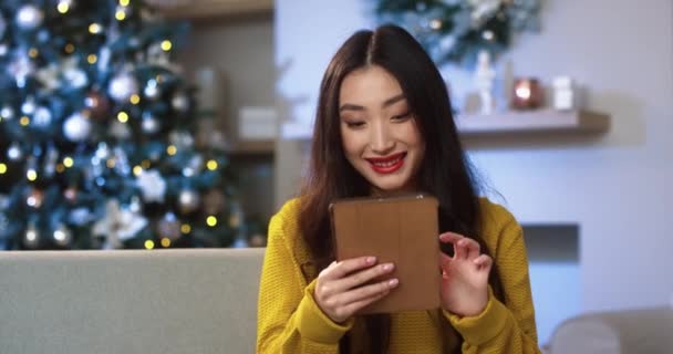 Portrét asijské radostné mladé krásné ženy v dobré náladě ve zdobeném moderním pokoji s vánoční strom klepání a rolování na tabletu a výběru vánoční dárky na Silvestra. Koncept slavnostní nálady