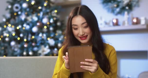 Asyalı mutlu genç bir kadının portresi süslü bir odada gülümsüyor ve yeni yıl ağacı tablete dokunuyor ve kayıyor ve Noel hediyelerini seçiyor. Şenlik havası. E-ticaret kavramı — Stok video