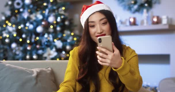 Close up portret młodej pięknej Azjatki szczęśliwy w urządzonym domu z świecących xmas drzewa wideo czatując na telefon komórkowy i machając ręką wysyłanie najlepszych widgetów na Boże Narodzenie. Koncepcja ducha Bożego Narodzenia — Wideo stockowe