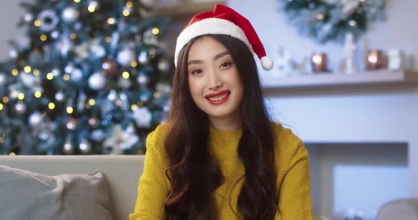 Close up portret wesołej młodej Azjatki w kapeluszu Mikołaja w świątecznym nastroju siedzi w urządzonym pokoju patrząc w aparat i uśmiechając się w Wigilię. Nowy Rok święto Zimowe święta Koncepcja Bożego Narodzenia — Wideo stockowe