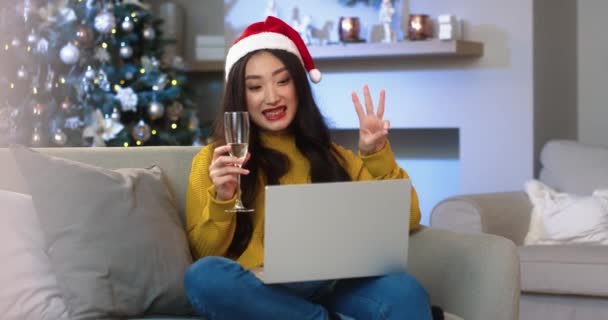 Glückliche junge schöne asiatische Frau mit Weihnachtsmütze in dekoriertem Raum mit leuchtenden Weihnachtsbaum videochatting auf Laptop hält ein Glas Champagner und tut Countdown für Silvesterfeier Feiertage Konzept — Stockvideo