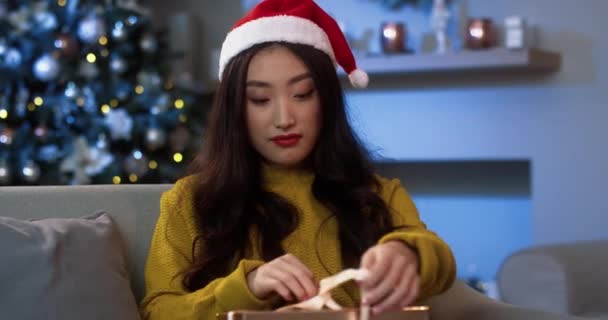 Close up retrato de alegre mulher jovem asiática abrindo xmas caixa de presente com rosto animado sentindo-se feliz e grato na véspera de Natal sentado em casa aconchegante perto de árvore de ano novo brilhante. Momentos mágicos — Vídeo de Stock
