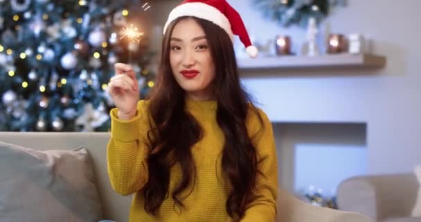Porträt einer glücklichen jungen Asiatin mit Weihnachtsmütze in festlicher Stimmung, die in einem geschmückten Raum mit Weihnachtsbaum sitzt und Wunderkerzen in der Hand hält. Neujahrsfeier. Konzept für Dezemberferien — Stockvideo