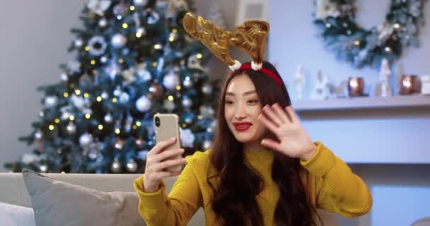 Młoda piękna, wesoła Azjatka w urządzonym pokoju ze świecącą choinką w świątecznym nastroju wideo na czacie przez telefon komórkowy i wysyłanie najlepszych widżetów na Boże Narodzenie koncepcja ducha Zamknij portret — Wideo stockowe