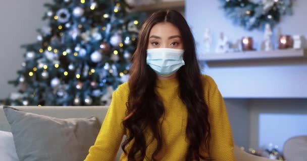 의료용 마스크를 착용하고 장식 된 방에 앉아 밝은 나무와 카메라를 보고 있는 아시아인젊은 여성의 사진을 클로즈업 해 보 세요. 크리스마스 장식. 격리 구역에서의 휴일 시즌. 유행병의 시대 — 비디오