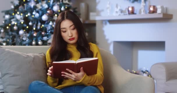 Retrato de asiática jovem bela fêmea sentada em quarto decorado em casa com árvore brilhante e ler livro sozinho na véspera de Natal. Decoração de Natal. Temporada de férias. Celebração de Ano Novo — Vídeo de Stock