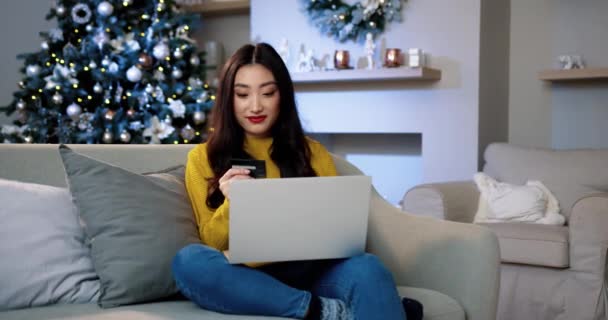 Felice donna asiatica sorridente seduta a casa in camera decorata la vigilia di Natale e l'acquisto di regali di Natale online sul computer portatile che paga con carta di credito. Shopping natalizio. Sconti natalizi. Concetto di commercio elettronico — Video Stock