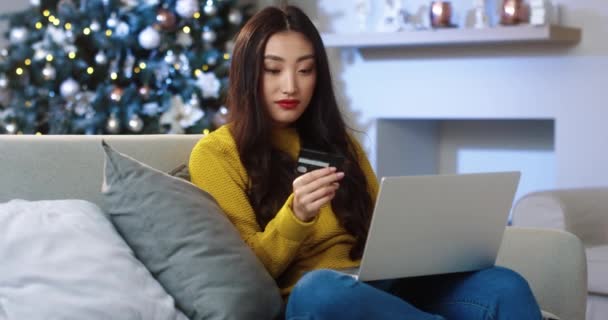Happy vzrušený asijské mladá žena sedí v zdobené místnosti na Vánoce a nákup vánoční dárky on-line na notebooku placení kreditní kartou na nákup na internetu. Nákupy. Koncept slev — Stock video
