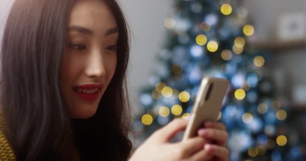 Primer plano retrato de feliz asiática bastante joven morena mensajes de texto femeninos y tocando en el teléfono móvil en el interior de casa cerca de árbol decorado. Mujer sonriendo y envía los mejores deseos con vacaciones en el teléfono celular — Vídeos de Stock