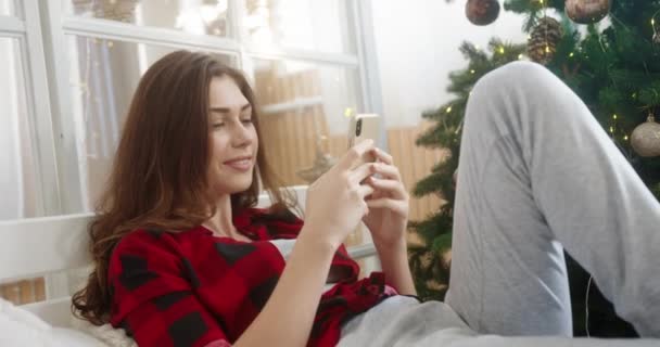 Close up of happy young beautiful Caucasian woman in bedroom near decorated christmas tree tapping and browsing on smartphone sending congratulation on Christmas Eve. Holidays concept — Stock Video