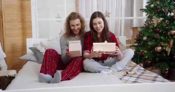 Close up of happy Caucasian young family couple sitting on bed in room at home on New Years Eve near xmas tree holding christmas gifts looking at camera and smiling. Xmas presents concept — Stock Video