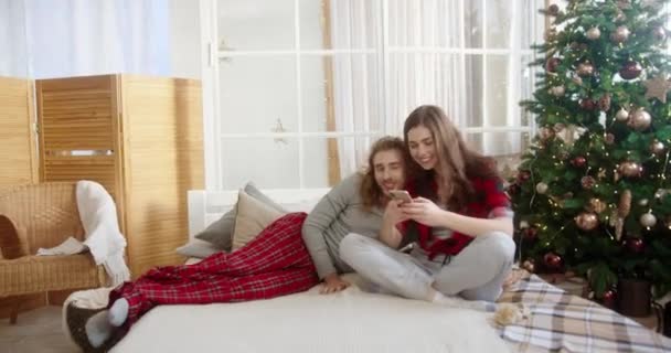 Blanke vrolijke jonge getrouwde echtpaar en echtgenoot zitten in de slaapkamer thuis door Kerstmis versierd gloeiende boom en typen op smartphone het verzenden van groeten met vakantie. Gezinsconcept — Stockvideo