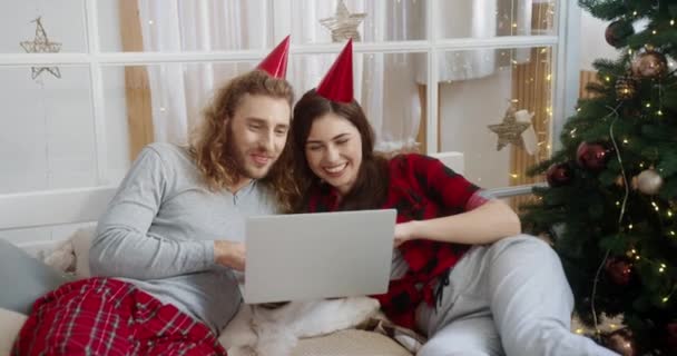 Großaufnahme des kaukasischen glücklichen jungen Familienpaares Frau und Mann, die im Schlafzimmer in der Nähe des geschmückten Weihnachtsbaums liegen und per Videoanruf am Laptop Neujahr feiern. Konzept für Weihnachtsferien — Stockvideo