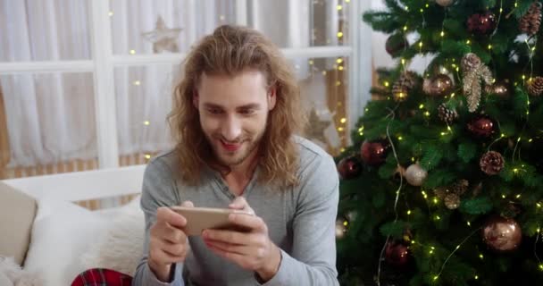 웃으며 웃고 있는 백인 남성이 밝게 빛나는 나무 근처의 집에서 침대에 앉아 있는 동안 핸드폰으로 타이핑하고 두드리는 즐거운 모습을 클로즈업 했다. 휴일 시즌 개념 — 비디오