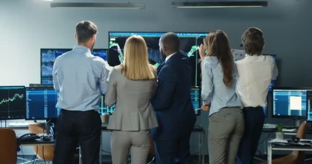 Zadní část mužských a ženských multietnických makléřů, kteří stojí v úřadu s monitory, obracejí se k fotoaparátu a oslavují úspěch. Veselí šťastní podnikatelé a podnikatelky tančí a smějí se po obchodování — Stock video