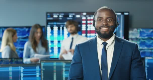 Portret van Afro-Amerikaanse man in pak en stropdas met hoofdtelefoon die glimlacht naar de camera in het beurskantoor. Handelsconcept. Professionele handelaar man in de kamer met monitoren en computers. Verkoopmanager. — Stockvideo