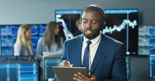 Agente joven afroamericano en auriculares hablando con el cliente y notando en la tableta. Hombre comerciante financiero comprar o vender acciones en gadget en la oficina de comercio. Mercado de cambio. Departamento de ventas. — Vídeo de stock