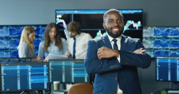 Porträtt av afroamerikansk man i kostym och slips leende till kameran och korsa händerna i börskontoret. Affärsidé. Professionell handlare man i rummet med bildskärmar. Försäljningschef. — Stockvideo