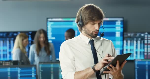 Joven corredor caucásico en auriculares hablando con el cliente y desplazándose en la tableta. Hombre comerciante financiero comprar o vender acciones en gadget en la oficina de comercio. Mercado de cambio. Departamento de ventas. — Vídeos de Stock