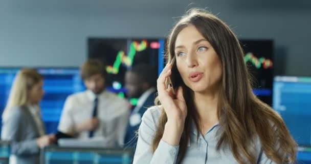 Vackra unga kaukasiska kvinna mäklare talar på mobiltelefon i börskontoret. Kvinnlig handlare talar på mobiltelefon i rummet med monitorer och kollegor. Affärskvinna chef. — Stockvideo