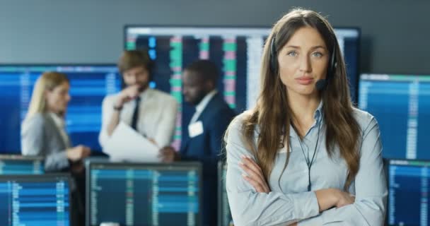 Retrato de hermosa mujer caucásica en auriculares mirando a la cámara y cruzando las manos en la oficina de bolsa. Concepto comercial. Comerciante femenina profesional en la habitación con monitores. Gerente de ventas. — Vídeos de Stock