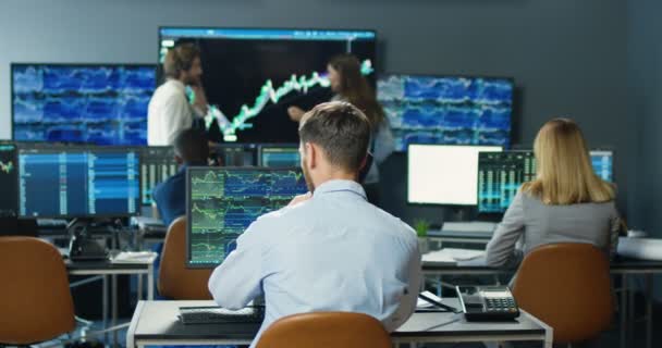 Arrière sur courtier homme caucasien assis à l'écran de l'ordinateur et répondre à l'appel téléphonique dans le bureau commercial. Vue arrière sur l'homme parlant au téléphone avec le client et travaillant avec les taux boursiers et les graphiques. — Video