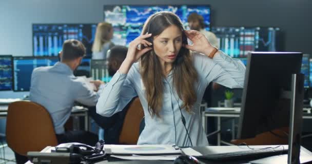 Kavkazská makléřka ve sluchátkách sedí u počítače v kanceláři a pracuje s dokumenty při prodeji nebo nákupu akcií. Žena se dívá na papíry s grafy a grafikou a mluví s klientem. — Stock video