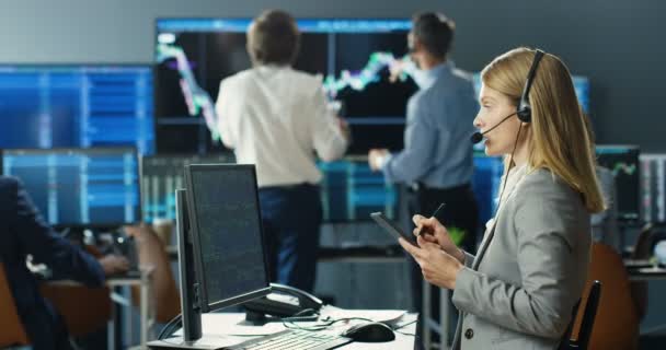 Mujer caucásica corredor de auriculares sentado delante del monitor de la computadora con gráficos, gráficos y tasas y hablar con el cliente. Observando en el dispositivo de tableta. Mujer comerciante hablando, comprando, vendiendo acciones. — Vídeos de Stock