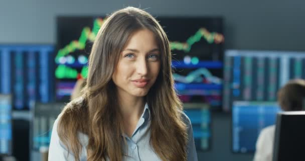 Portrait de jeune femme commerçante Caucasienne belle avec les cheveux longs regardant la caméra et souriant dans le bureau de négoce de bourse. Moniteurs avec graphiques, graphiques et chiffres en arrière-plan. — Video