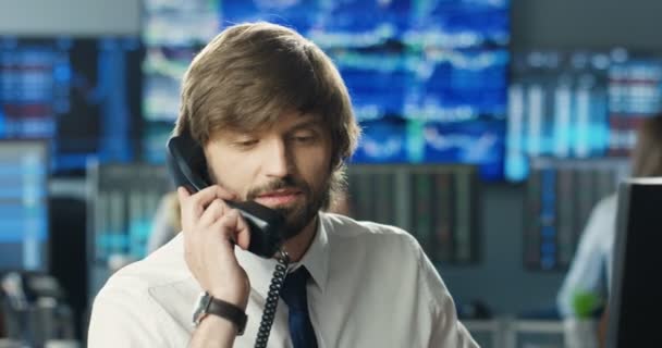 Close up de belo jovem caucasiano corretor trabalhando no monitor do computador e atender telefonema. Comerciante masculino do mercado de ações falando por telefone no escritório de negociação. Um negócio bem sucedido. Dia de trabalho. — Vídeo de Stock