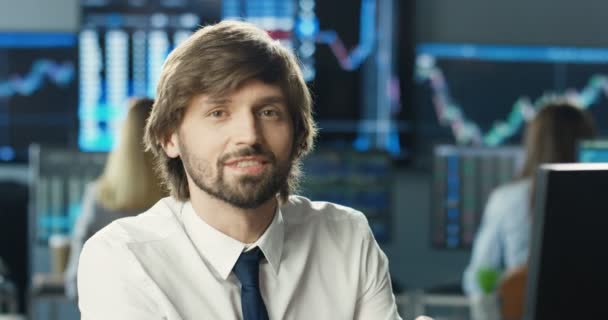Portret van de jonge blanke knappe man handelaar in stropdas en met baard kijkend naar de camera en glimlachend in het handelskantoor van de beurs aandelenmarkt. Monitoren met grafieken en nummers op de achtergrond. — Stockvideo