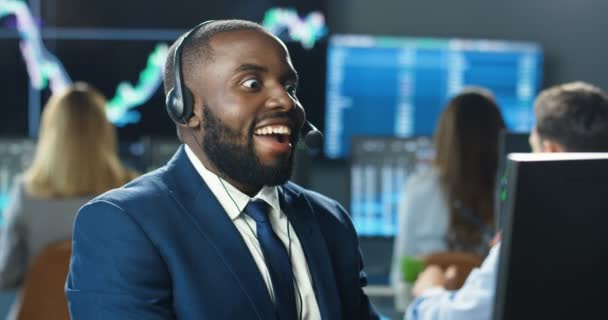 Afro-americano guapo joven comerciante masculino en auriculares que trabajan en el ordenador monior y que tienen un gran resultado en el comercio. Hombre corredor de conseguir un buen resultado y se sorprende. Feliz noticia en la oficina de oficios. — Vídeos de Stock