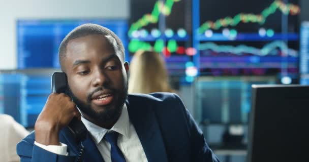 Närbild av stilig afroamerikansk man mäklare som jobbar vid datorn och svarar i telefon. Man börshandlare talar i telefon på handelskontoret. Telefonaffärssamtal. — Stockvideo