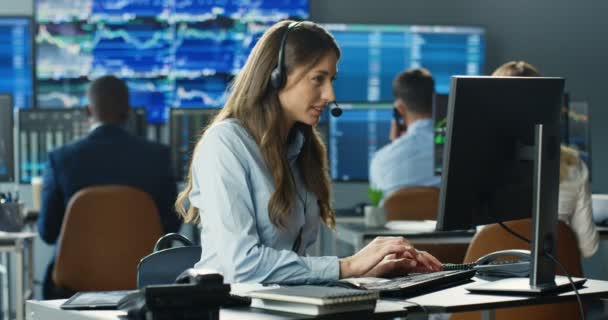 Young Caucasian pretty woman trader in headset working at monitor of computer in trading office and talking with client. Female broker typing on keyboard and speaking with customer. Sales call center. — Stock Video