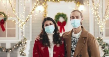 Yeni yıl arifesinde kar yağışı sırasında şehirde sokakta duran, tıbbi maskeli, mutlu, beyaz, evli bir çiftin portresi. Karantina Xmas konseptinde Noel zamanı şenlik havasında olan kadın ve erkek