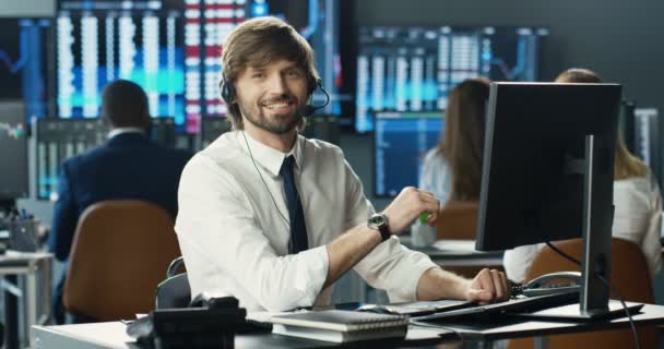 Retrato joven caucásico hombre guapo comerciante en auriculares trabajando en la computadora, mirando a la cámara y sonriendo en la oficina de comercio. Bolsa de valores. Feliz alegre macho exitoso corredor con sonrisa. Centro de llamadas. — Vídeo de stock